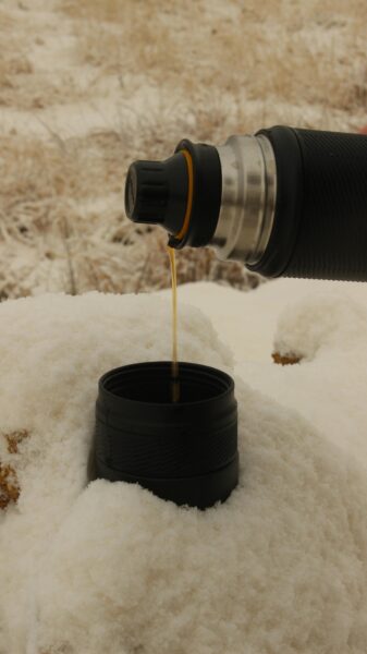 A close-up of a hot drink being poured from a thermos into a cup in a snowy setting.