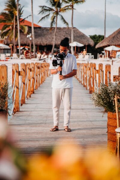A professional filmmaker with a camera on a gimbal, filming at a tropical resort pier during summer.
