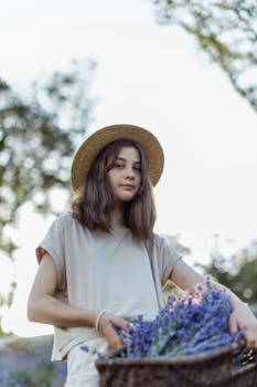 A young girl wearing a straw hat holds a basket filled with lavender flowers outdoors.