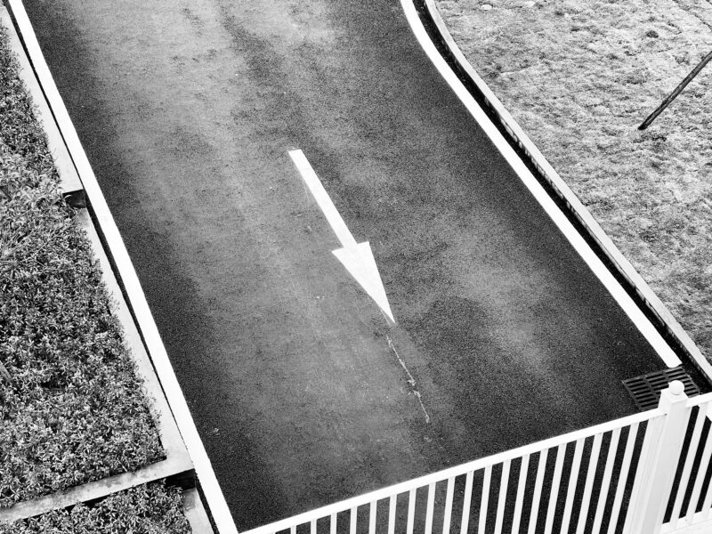 Black and white overhead shot of a road with an arrow, bordered by grass and fence.