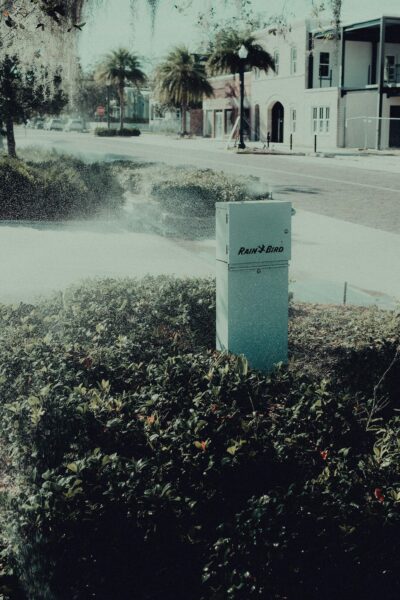 Sprinkler system in a green urban area with surrounding buildings on a sunny day.