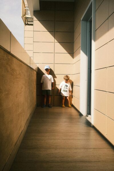 Two young boys stand on a sunlit urban balcony, casting shadows against modern architecture