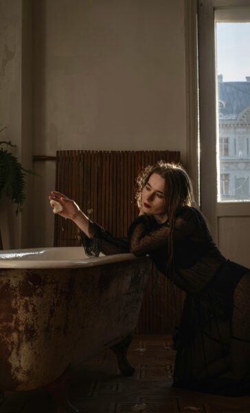 A woman in a black dress leans on a vintage bathtub, creating an artistic and dramatic indoor scene.