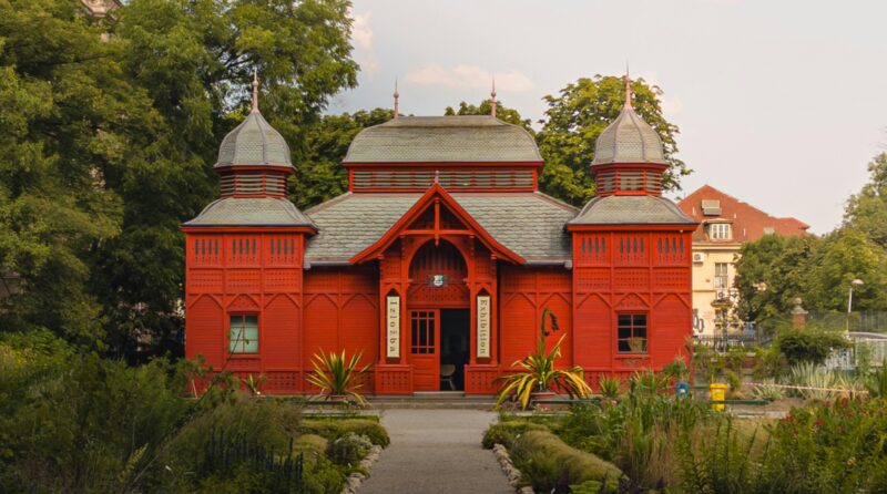 Historic red wooden pavilion in Zagreb Botanical Garden, surrounded by lush greenery.
