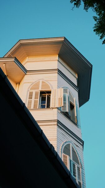 A Victorian-style tower with open shutters against a clear blue sky, reflecting warm afternoon sunlight.