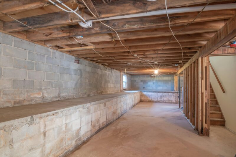 Empty basement with exposed wooden beams, concrete walls, and stairs.