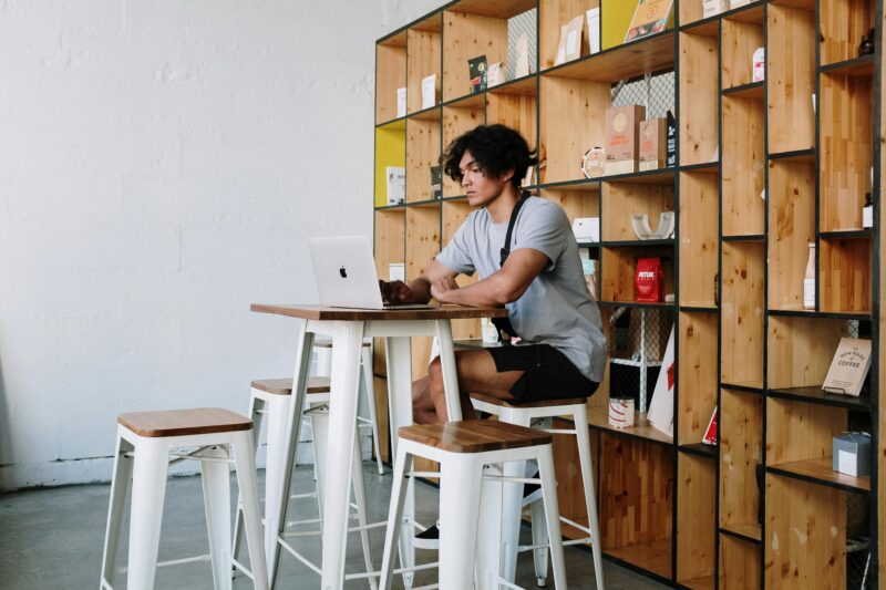 A young man sits in a loft cafe working on a laptop, surrounded by wooden decor and shelving.