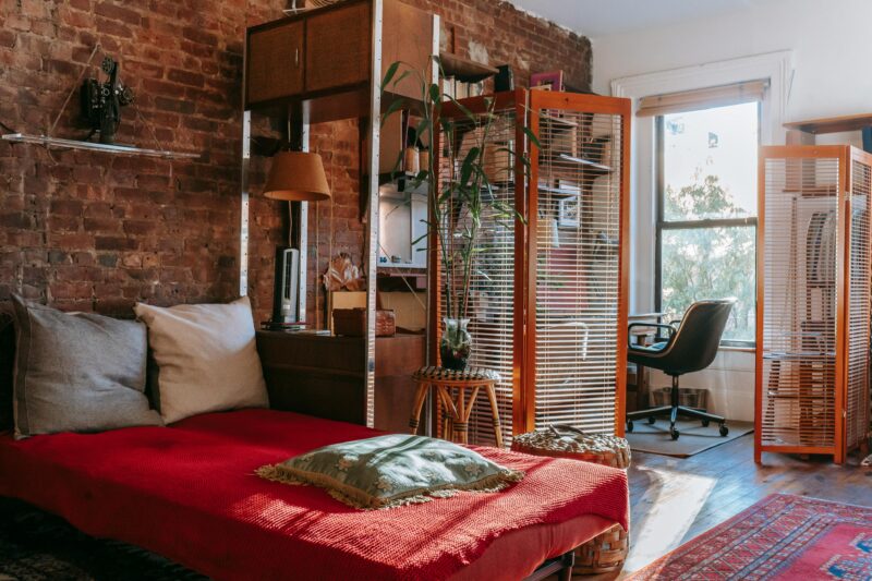 Comfortable bed with red cover and pillows placed near brick wall in cozy room with isolated workspace decorated with houseplants near window.