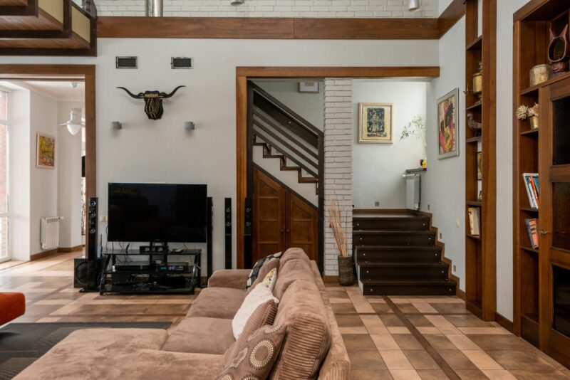 Interior of modern apartment with comfortable couch and TV placed near decorated wall with stairway at wooden shelves at home