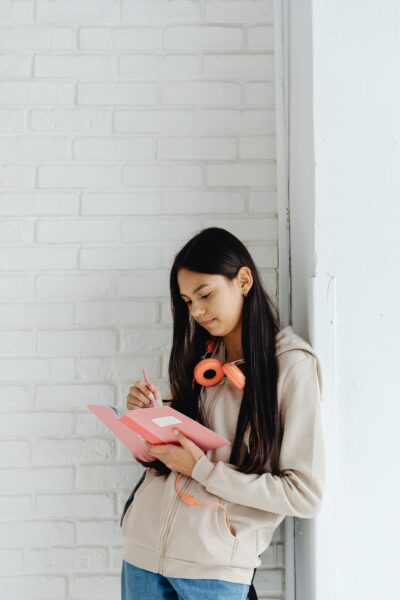 Teen girl writing in a notebook, leaning against a white brick wall, with headphones around her neck.