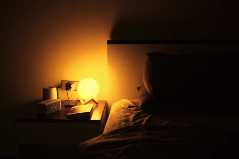 A night stand with a lamp on it next to a bed.