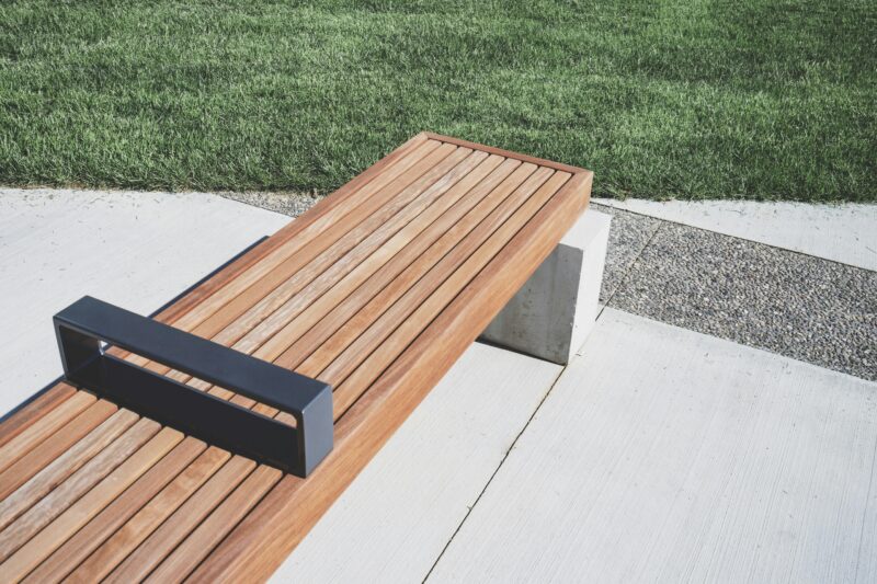 A minimalist wooden bench placed on a concrete path beside a green lawn in daylight.