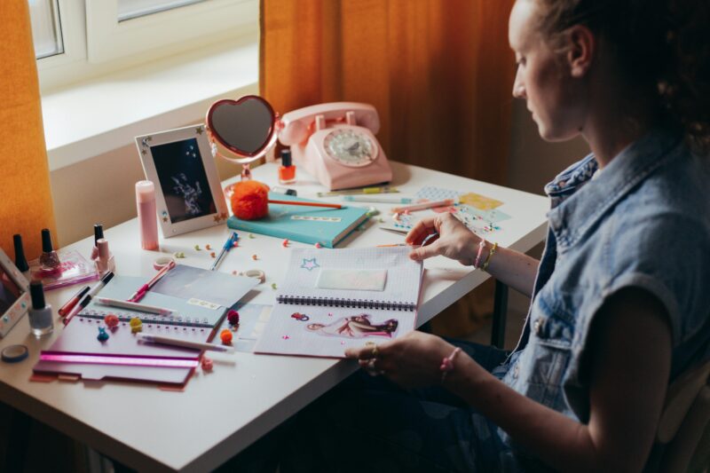 A woman sits at a cluttered desk working on a creative journal, surrounded by stationery and decorative items.
