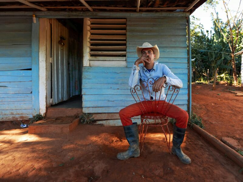 A cowboy in red pants and boots sits relaxed outside a rustic wooden house, exuding rural charm.