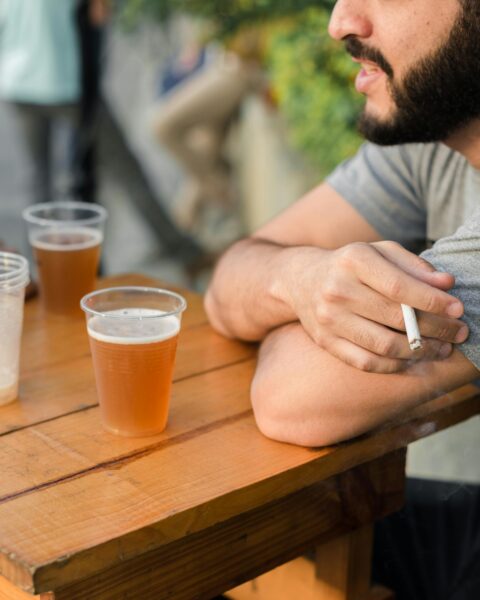 Adult man with a cigarette enjoying a beer at an outdoor wooden table, capturing casual leisure time.