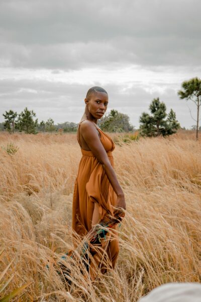 A stylish woman poses elegantly in a golden field under a cloudy sky.