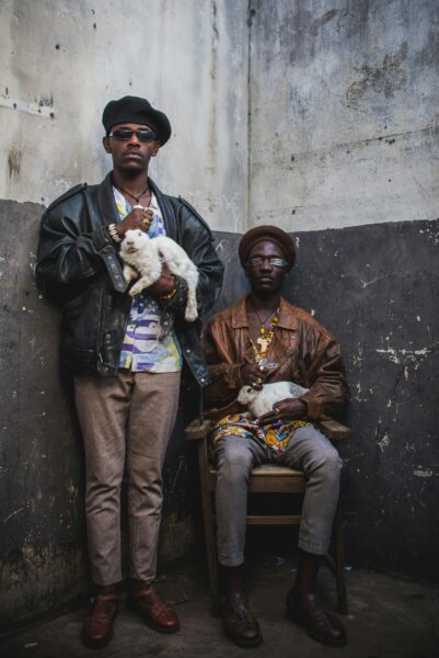 Two fashionable men pose with cats in an urban environment, showcasing unique style.