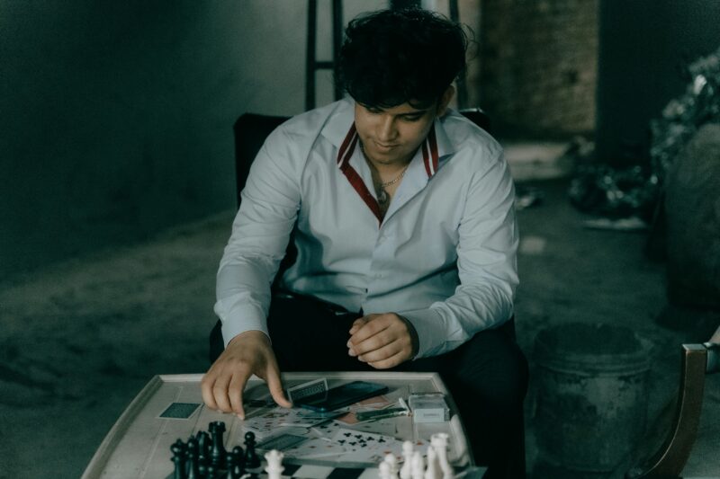 A man in a button-up shirt thinks about his chess move at a wooden table in a dimly lit room.