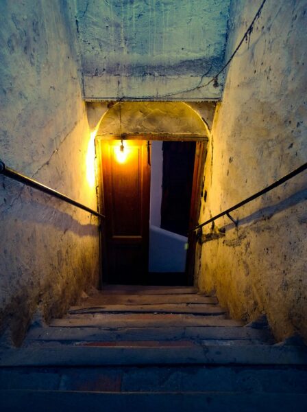 A narrow stone staircase illuminated by a single bulb leading to a closed wooden door.