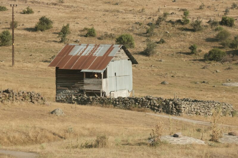 An old, weathered shed stands in an arid rural landscape, surrounded by stone walls.