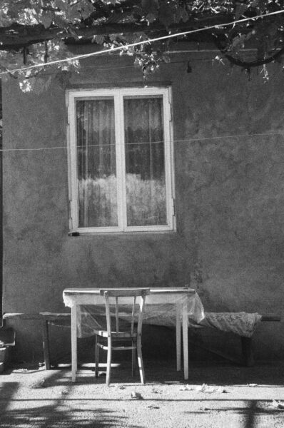 A black and white image of a rustic outdoor space featuring a window, table, and chair.