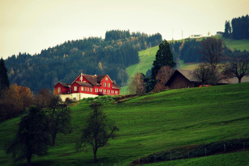 A red farmhouse nestled in a misty, green countryside with rolling hills and distant forests.