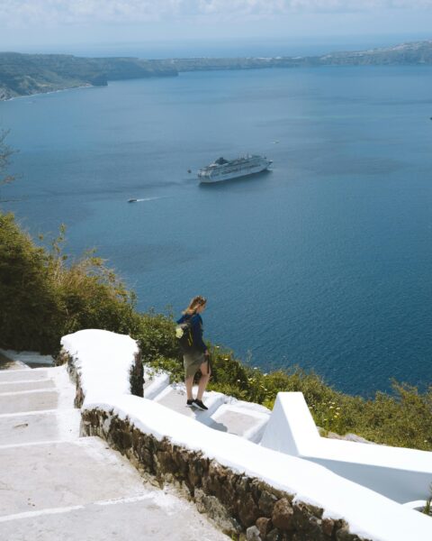 A woman walks down white stairs with a cruise ship in view, overlooking the Aegean Sea in Santorini, Greece.