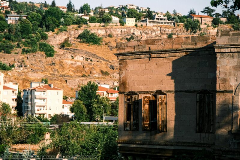 A rustic stone building with shutters set against a sunny hillside and residential area.