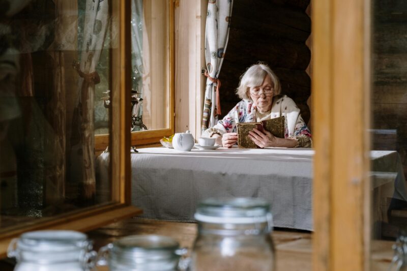Senior woman reads by the window in a cozy cabin, enjoying a cup of tea with a teapot on the table.