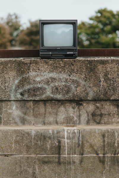 Black old fashioned TV placed on shabby stone fencing with graffiti in city on street with green trees.