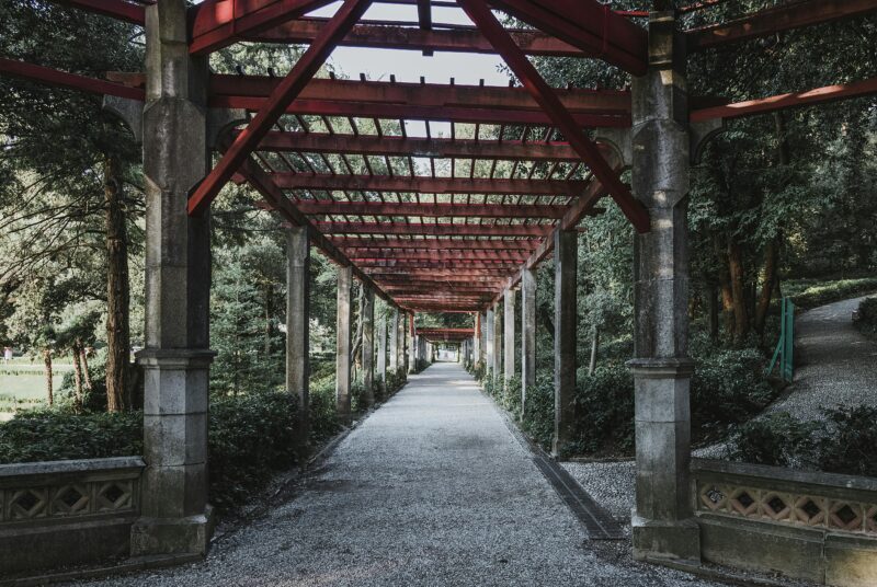 View of a rustic pergola structure along a path in a lush garden, surrounded by greenery.