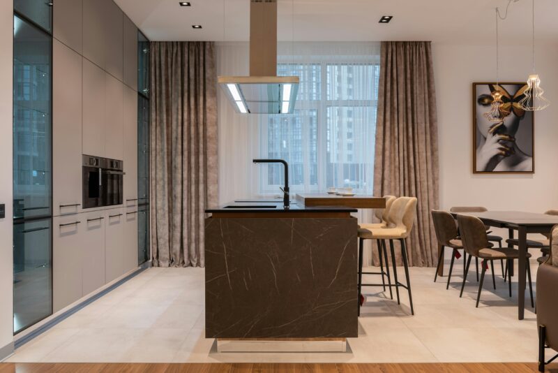 Interior of contemporary room with counter and chairs near window with curtains near table and kitchenware and cabinet