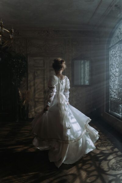 A woman in a flowing white dress stands in a vintage room with ornate details, exuding elegance and mystery.