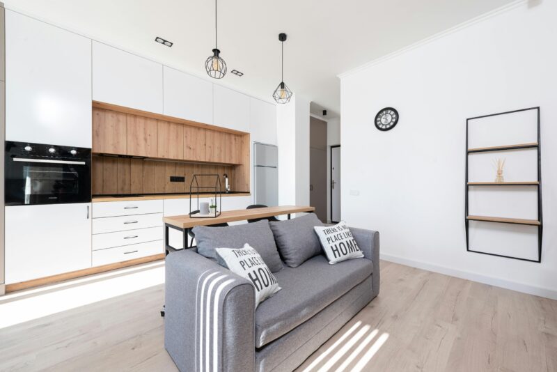Interior of bright modern room with open kitchen with cupboards and oven next to couch with pillows and table.