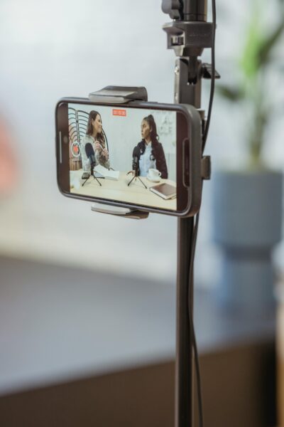 Young diverse women sitting at table with microphones and gadgets and talking while recording podcast on smartphone placed on tripod in studio.