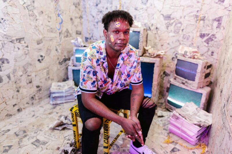 A man in a colorful shirt with face stickers sits in a vintage-style room full of old TVs and newspapers.