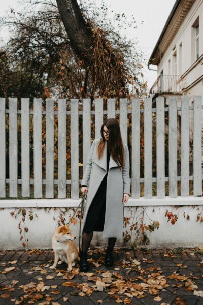 A woman with glasses stands with a Shiba Inu dog on a leash against an autumn background.