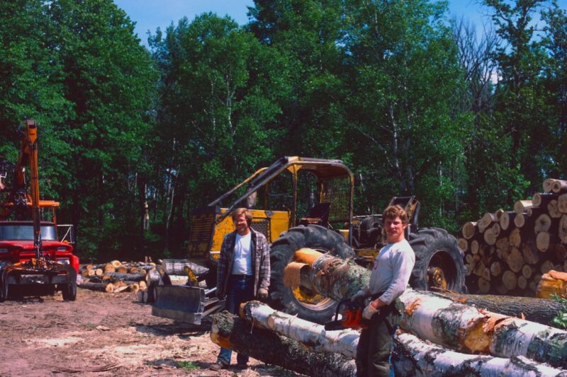 Two men standing near cut logs beside trees.