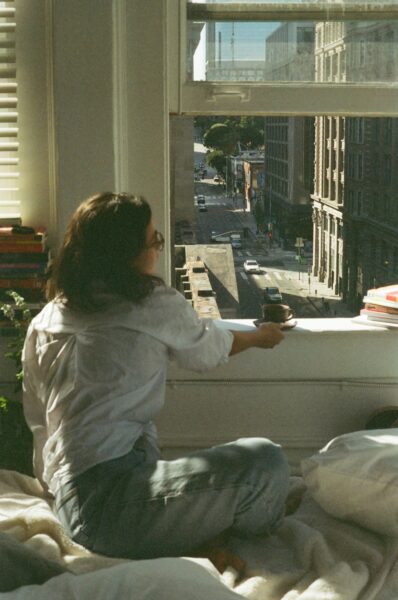 Woman in white hoodie sitting on bed enjoying espresso.