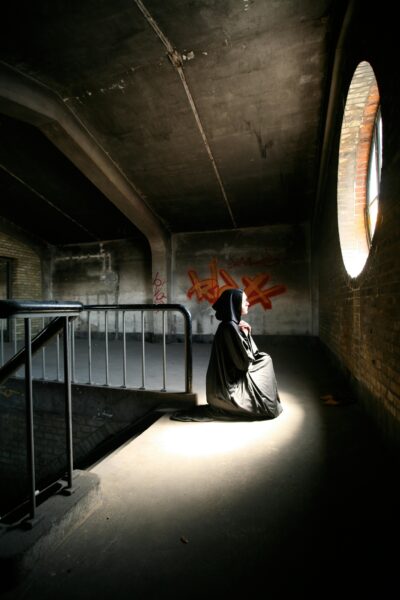 A solitary figure in a black cloak kneels in a dimly lit attic space, creating an eerie atmosphere.