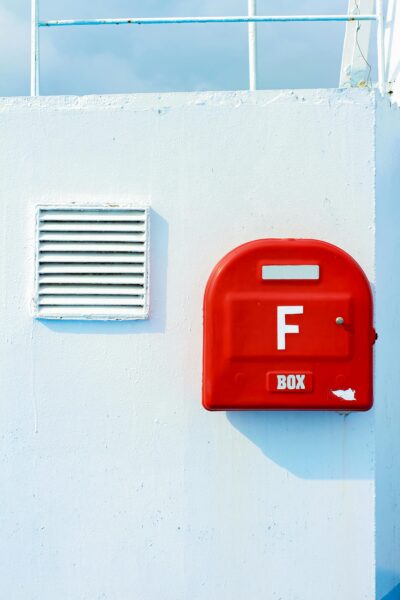 Red mailbox mounted on a white wall outdoors, casting a shadow under blue sky.
