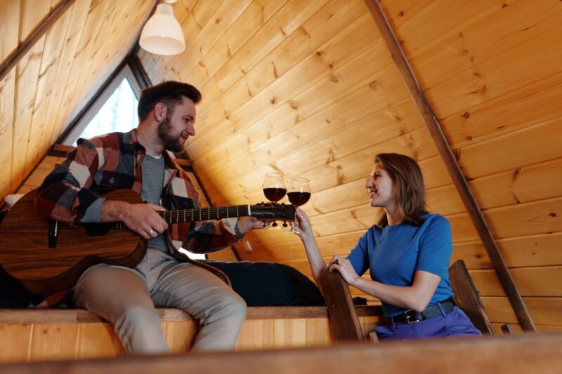 A couple enjoys wine and guitar music in a cozy wooden cabin, creating a warm and intimate atmosphere.