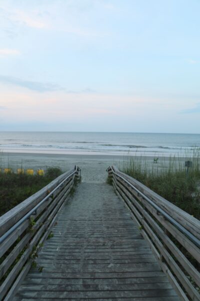 A tranquil beach scene with a wooden pathway leading to the ocean under a clear sky.