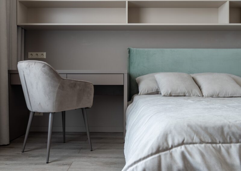 Interior of modern light bedroom with pillows and blanket on bed near table with chair under shelves next to curtains