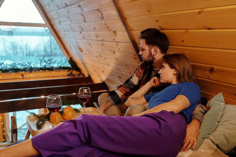 Relaxed couple lounging in a wooden cabin, sharing wine with scenic lake view.